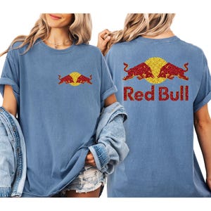 May include: A light blue t-shirt featuring the Red Bull logo on the front and back. The logo includes two red bulls facing each other with a yellow circle in the center. The words "Red Bull" are printed in red glitter.