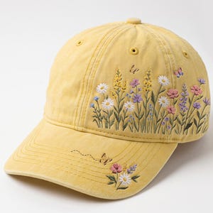 May include: A yellow baseball cap featuring detailed embroidery of wildflowers, including white daisies, purple and yellow stalks, and small pink blossoms. A small butterfly is embroidered on the brim and side, with a dotted flight path trail.