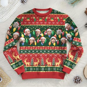 Peut inclure: Un pull de Noël festif rouge, vert et blanc. Le pull présente un motif de visages portant des bonnets de Père Noël et des guirlandes lumineuses. Les manches ont des motifs de rennes et de sapins.