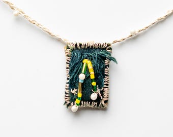 May include: A rectangular pendant on a light tan cord featuring a dark green fabric base with frayed edges. The pendant is decorated with a curved line of yellow and green beads, small white pearls, and metallic accents stitched onto the fabric.
