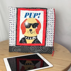 May include: A quilted tablet case featuring a cartoon dog wearing sunglasses and a studded collar, with the word "PUP!" above. The case has a red, black, and white color scheme with a zipper closure and a patterned design.