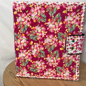 May include: A quilted journal cover with a vibrant floral pattern in shades of pink, yellow, and green on a deep red background. The cover has a white border and a matching closure with green buttons. The journal cover is sitting on a wooden table.