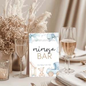 Könnte beinhalten: Ein Mimosa Bar-Schild mit Text in Schwarz und Gold, mit einer Illustration eines Kinderwagens und eines Teddybären. Das Schild steht auf einem Tisch mit Sektgläsern und Blumengestecken. Der Text lautet: "Gießen Sie das Sprudelnde ein, spritzen Sie etwas Saft, fügen Sie etwas Obst hinzu - jetzt nippen und loslassen!"
