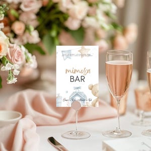 Könnte beinhalten: Ein Mimosa Bar-Schild mit goldfarbenen Buchstaben, gehalten von einem transparenten Glasständer. Das Schild hat eine Aquarellillustration eines Teddybären und eines Kinderwagens. Zwei Sektgläser mit einem rosa Getränk sind im Vordergrund.