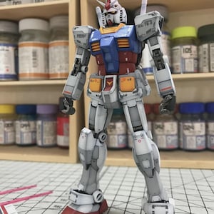 以下が含まれることがあります： 白、青、赤、黄色のガンダムロボットの詳細なモデル。モデルは「ArrowModelBuild」のテキストが見えるグリッド面に立っています。ロボットは複雑な機械的詳細を持っています。