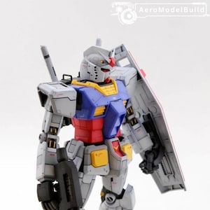 May include: A detailed model of a Gundam robot, primarily gray with blue, red, and yellow accents. It holds a black rifle and a shield. The model is standing upright, showcasing its intricate design and mechanical details.
