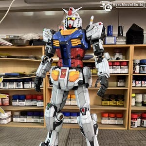 May include: A detailed model of a Gundam robot, featuring a color scheme of white, blue, red, yellow, and gray. The model has intricate panel lines and markings, with visible mechanical details. The robot is standing upright, showcasing its articulated limbs and weaponry.