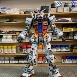 K&ouml;nnte beinhalten: Ein detailliertes Modell des RX-78-2 Gundam, ein wei&szlig;er, roter, blauer und gelber Mecha-Roboter, der aufrecht steht. Ein kleineres rotes und blaues Raumschiffmodell befindet sich davor. Das Modell steht auf einer Schneidematte mit einem Lineal.