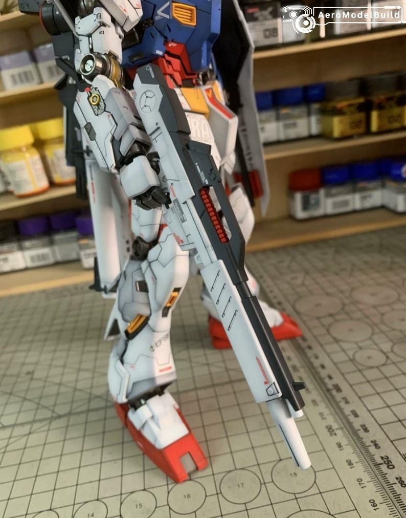 K&ouml;nnte beinhalten: Detailliertes Modell eines Roboters, haupts&auml;chlich wei&szlig; mit roten, blauen und gelben Akzenten. Der Roboter h&auml;lt eine gro&szlig;e, schwarz-wei&szlig;e Waffe. Das Modell ist teilweise montiert und zeigt komplizierte mechanische Details. Der Text "AeroModel Build" ist sichtbar.