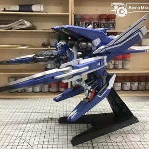 May include: A detailed model of a blue and white mecha robot, featuring intricate wing-like structures and a black base. The model is displayed on a grid-patterned surface, with shelves and paint supplies in the background.