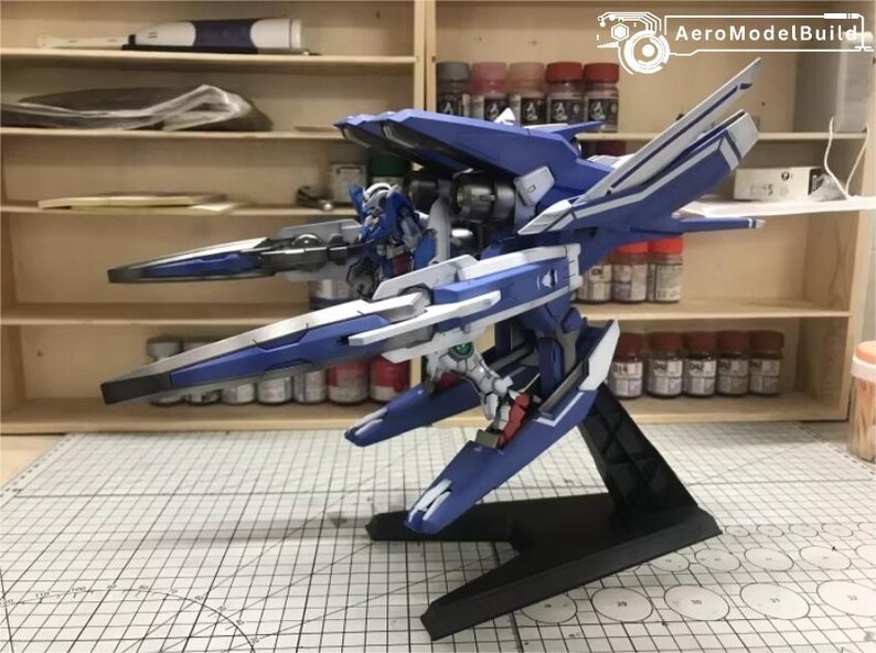 May include: A detailed model of a blue and white mecha robot, featuring intricate wing-like structures and a black base. The model is displayed on a grid-patterned surface, with shelves and paint supplies in the background.