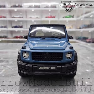 May include: A blue model of a Mercedes-Benz G-Class SUV. The model has black accents, a black grille with the Mercedes-Benz logo, and the AMG logo on the front bumper. The model is displayed in front of a shelf with other model cars.