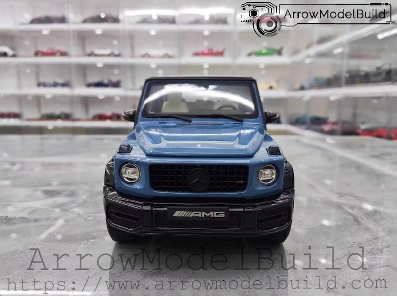 May include: A blue model of a Mercedes-Benz G-Class SUV. The model has black accents, a black grille with the Mercedes-Benz logo, and the AMG logo on the front bumper. The model is displayed in front of a shelf with other model cars.