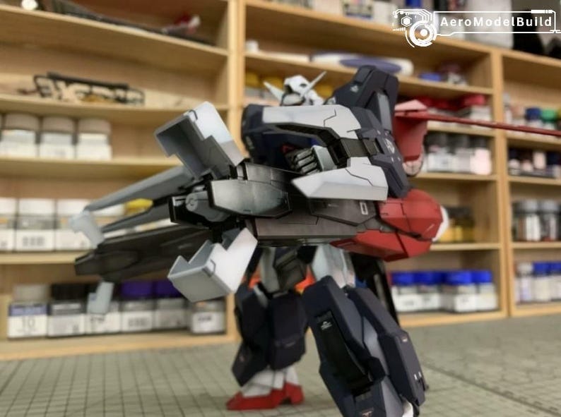 Puede incluir: Modelo detallado de un robot mecha, con una combinaci&oacute;n de colores blanco, azul marino, rojo y gris. El robot tiene extremidades articuladas y est&aacute; posado en una postura din&aacute;mica. El logotipo de AeroModelBuild es visible en la esquina superior derecha.
