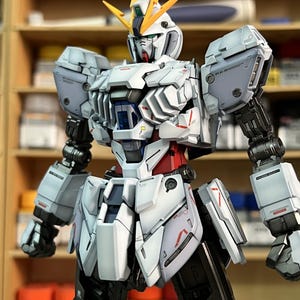 May include: A detailed model of a white, gray, red, and black robot with yellow accents. The robot has a complex design with articulated joints and panels. The image includes the text "ArrowModelBuild" in the upper right and lower left corners.