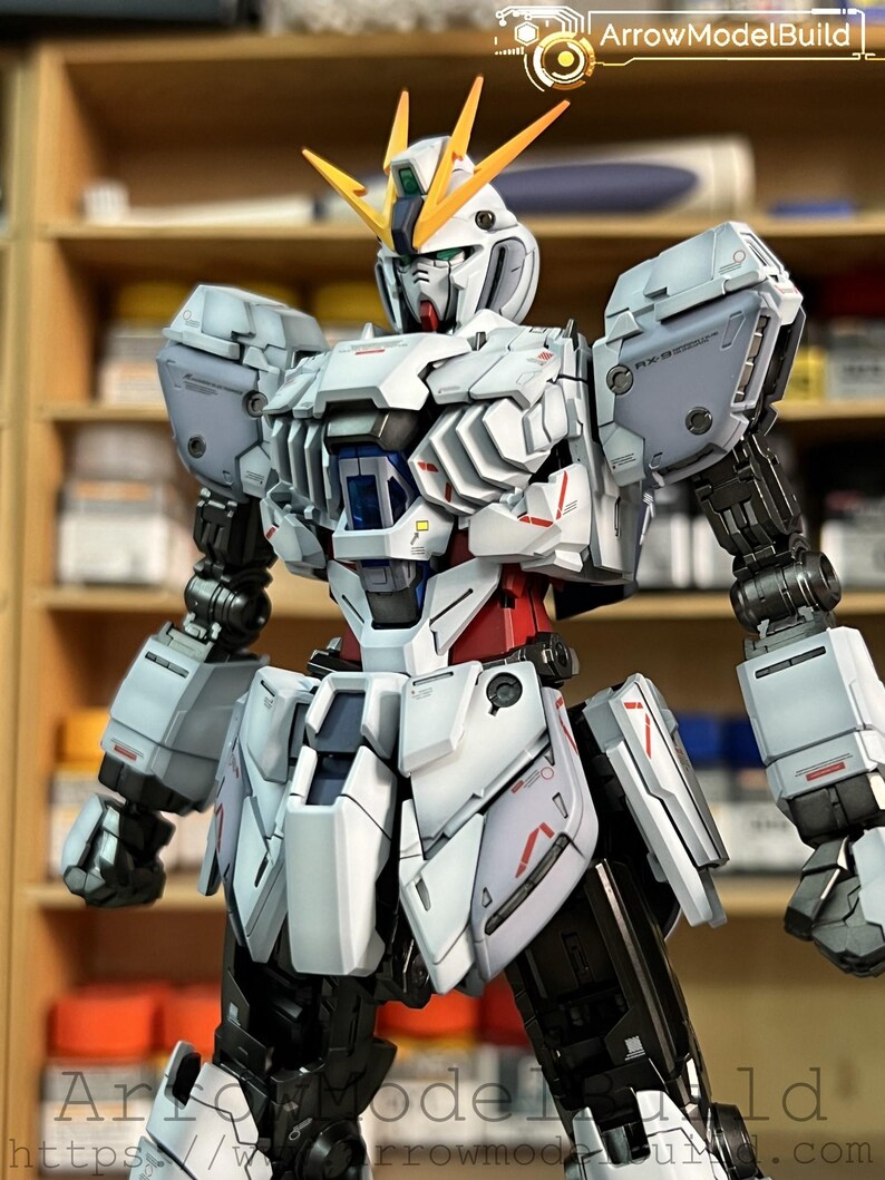 May include: A detailed model of a white, gray, red, and black robot with yellow accents. The robot has a complex design with articulated joints and panels. The image includes the text "ArrowModelBuild" in the upper right and lower left corners.