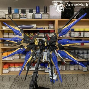 Puede incluir: Maqueta detallada de un robot Gundam, con una combinaci&oacute;n de colores negro, dorado, azul y blanco. El modelo presenta grandes alas estilizadas y detalles intrincados. El fondo muestra estanter&iacute;as con varias pinturas y herramientas, sugiriendo un tema de hobby o modelismo.