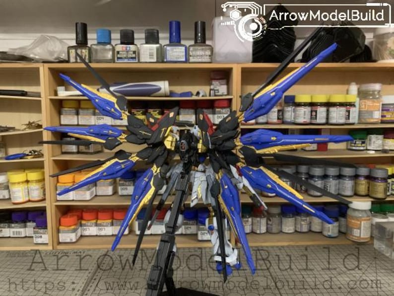 Puede incluir: Maqueta detallada de un robot Gundam, con una combinaci&oacute;n de colores negro, dorado, azul y blanco. El modelo presenta grandes alas estilizadas y detalles intrincados. El fondo muestra estanter&iacute;as con varias pinturas y herramientas, sugiriendo un tema de hobby o modelismo.