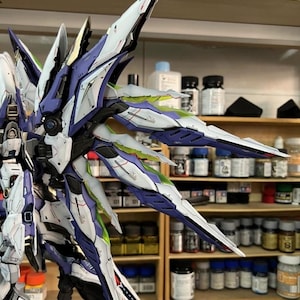 May include: A highly detailed model robot with large, stylized wings. The model is predominantly white, with blue, purple, and green accents. The wings have sharp, angular designs. The model is set against a backdrop of paint bottles and shelves.
