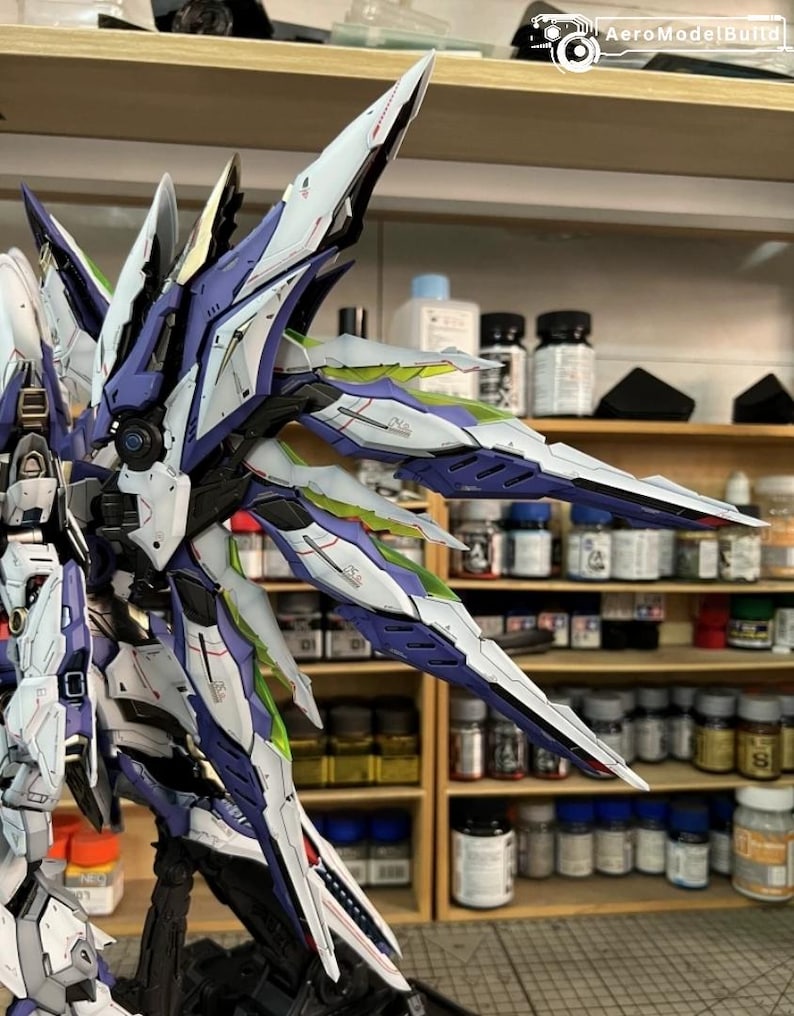 May include: A highly detailed model robot with large, stylized wings. The model is predominantly white, with blue, purple, and green accents. The wings have sharp, angular designs. The model is set against a backdrop of paint bottles and shelves.