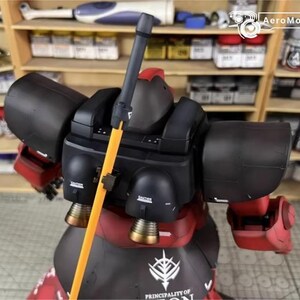 May include: A detailed model of a red and black robot with an orange pole. The robot has large black shoulder pads and a red torso. The model has gold-colored thrusters and the text "PRINCIPALITY OF ZEON" on the base.