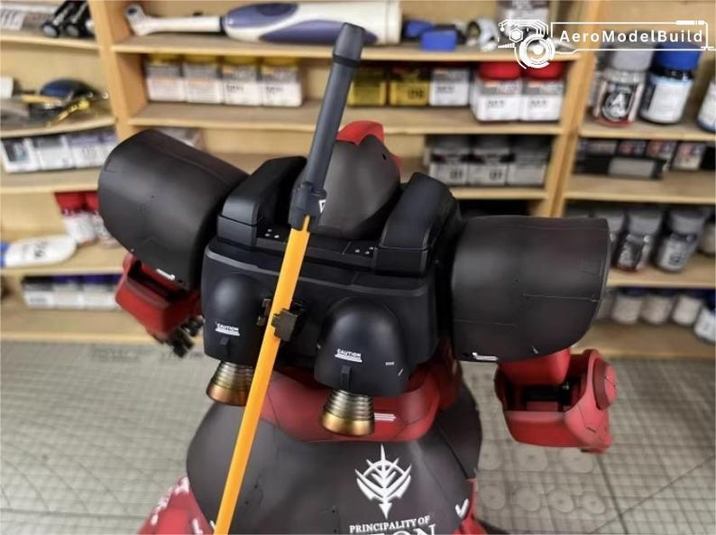 May include: A detailed model of a red and black robot with an orange pole. The robot has large black shoulder pads and a red torso. The model has gold-colored thrusters and the text "PRINCIPALITY OF ZEON" on the base.