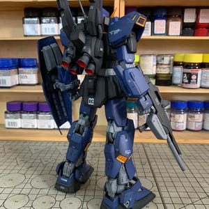 May include: A detailed model kit of a mecha robot, primarily in navy blue and black with orange and gray accents. The robot is posed with a gun and shield, showcasing intricate mechanical details. The model is on a grid surface.
