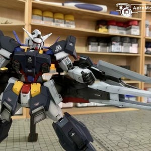 Puede incluir: Modelo detallado de un robot Gundam, con una combinaci&oacute;n de colores azul marino, blanco, amarillo y rojo. El modelo est&aacute; posado con los brazos extendidos, mostrando intrincados detalles mec&aacute;nicos. El fondo incluye estantes con suministros de pintura.