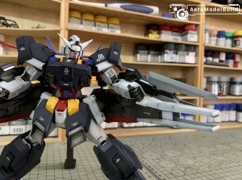 Puede incluir: Modelo detallado de un robot Gundam, con una combinaci&oacute;n de colores azul marino, blanco, amarillo y rojo. El modelo est&aacute; posado con los brazos extendidos, mostrando intrincados detalles mec&aacute;nicos. El fondo incluye estantes con suministros de pintura.