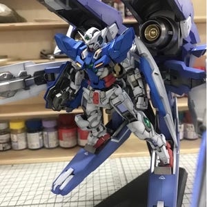 May include: A detailed model of a Gundam robot, primarily white, blue, and red. The model has intricate mechanical details, including wings and weapons. The background shows a workshop setting with paint bottles and shelves.