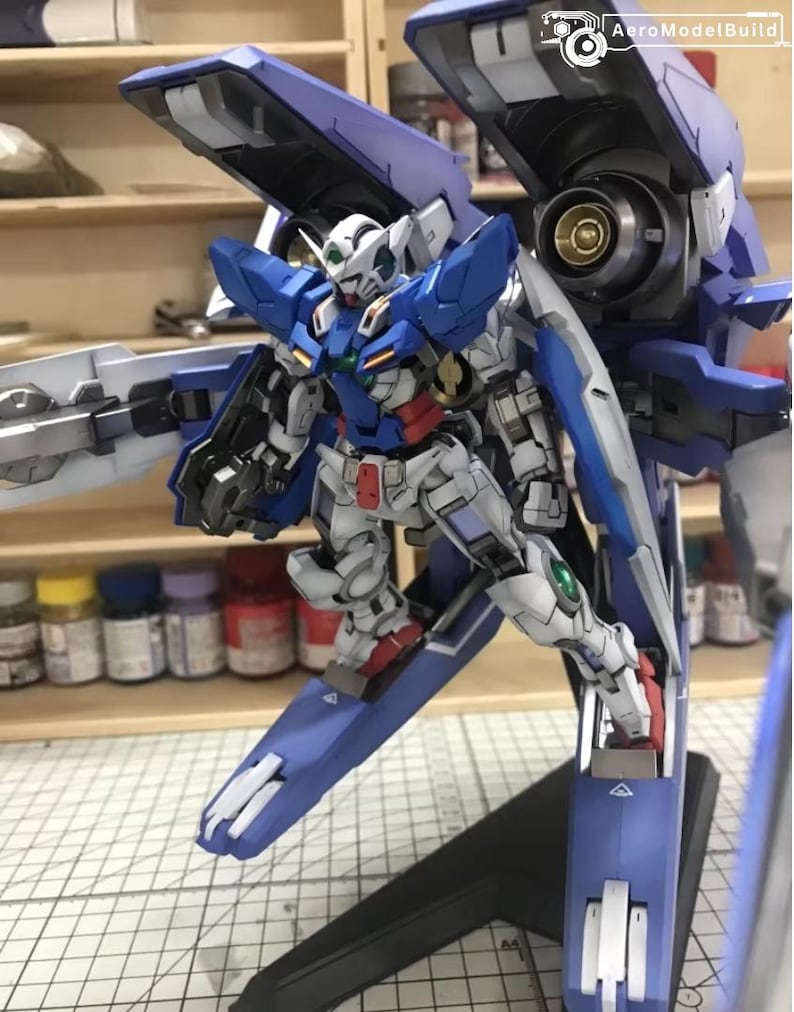 May include: A detailed model of a Gundam robot, primarily white, blue, and red. The model has intricate mechanical details, including wings and weapons. The background shows a workshop setting with paint bottles and shelves.