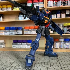 May include: A detailed, poseable model kit of a mecha robot, primarily navy blue with orange and yellow accents. The robot is holding a large black rifle and is standing on a grid-patterned surface. The model is surrounded by shelves of paint jars.