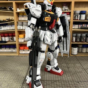 May include: A detailed model of a Gundam robot, predominantly white with black, red, and yellow accents. The model is holding a black rifle and has a shield. The model is standing in front of a shelf with various paint bottles and tools.