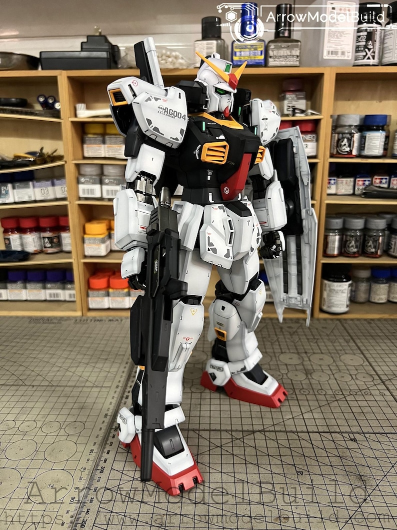 May include: A detailed model of a Gundam robot, predominantly white with black, red, and yellow accents. The model is holding a black rifle and has a shield. The model is standing in front of a shelf with various paint bottles and tools.