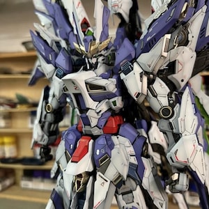 May include: A highly detailed mecha robot model, predominantly white, purple, red, and gold. The model features intricate mechanical detailing, large wings, and a futuristic design. The image highlights the model's complex construction and various components.