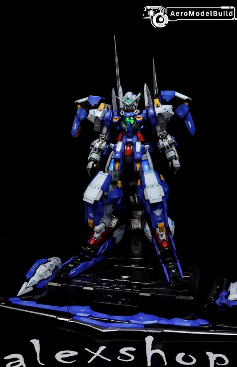 May include: A detailed model of a futuristic robot, primarily blue and white, with red and yellow accents. The robot is standing on a black base. The image includes the text "AeroModelBuild" and "alexshop".