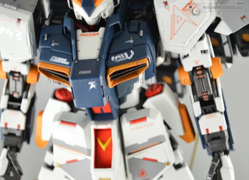 May include: Close-up of a detailed, articulated robot model. The model features a color scheme of white, navy blue, and orange. The robot has intricate mechanical details, including panels, vents, and markings. The model is a collectible item.