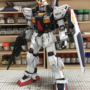 Op de afbeelding: Een gedetailleerd model van een witte, zwarte en rode RX-178 Gundam-robot met een schild en een geweer. Het model heeft gele en oranje accenten en staat op een rasteroppervlak. Het model is een gedetailleerde weergave van een mecha-robot.