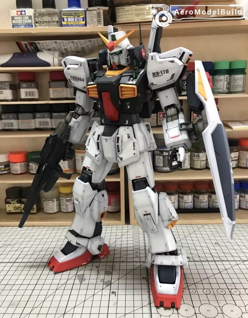 Op de afbeelding: Een gedetailleerd model van een witte, zwarte en rode RX-178 Gundam-robot met een schild en een geweer. Het model heeft gele en oranje accenten en staat op een rasteroppervlak. Het model is een gedetailleerde weergave van een mecha-robot.
