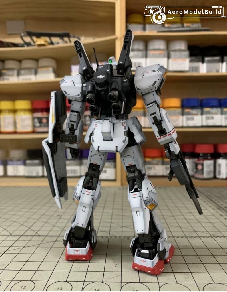 Pode incluir: Um modelo de rob&ocirc; detalhado e montado, principalmente branco com detalhes cinza e preto. O rob&ocirc; apresenta membros articulados, um escudo e uma arma. O modelo est&aacute; situado em um fundo de potes de tinta e prateleiras, sugerindo um tema de hobby ou modelagem.