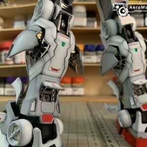 K&ouml;nnte beinhalten: Zwei detaillierte, teilweise montierte Modellroboterbeine. Die Beine sind haupts&auml;chlich wei&szlig; mit grauen und roten Akzenten und weisen komplizierte mechanische Details auf. Das Bild enth&auml;lt das AeroModelBuild-Logo in der oberen rechten Ecke.