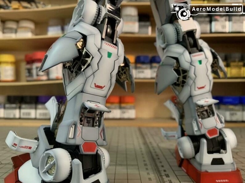 K&ouml;nnte beinhalten: Zwei detaillierte, teilweise montierte Modellroboterbeine. Die Beine sind haupts&auml;chlich wei&szlig; mit grauen und roten Akzenten und weisen komplizierte mechanische Details auf. Das Bild enth&auml;lt das AeroModelBuild-Logo in der oberen rechten Ecke.