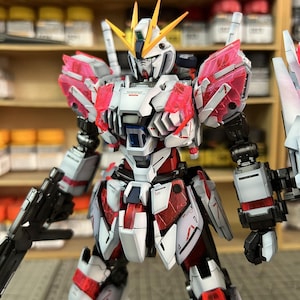 May include: A detailed model of a mecha robot, primarily white with red and pink accents. The robot has a complex design with multiple panels, weapons, and a yellow and white head. The image includes the text "ArrowModelBuild" and a website address.