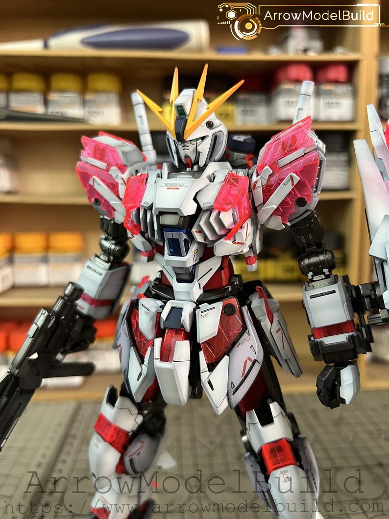 May include: A detailed model of a mecha robot, primarily white with red and pink accents. The robot has a complex design with multiple panels, weapons, and a yellow and white head. The image includes the text "ArrowModelBuild" and a website address.