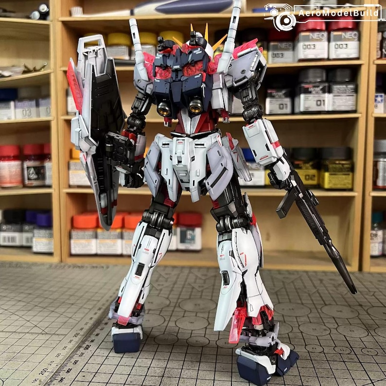 May include: A detailed model of a robot with a white, red, and black color scheme. The robot has a shield, a rifle, and multiple antennas. The model is standing on a surface with a ruler and is in front of a shelf with paint supplies.