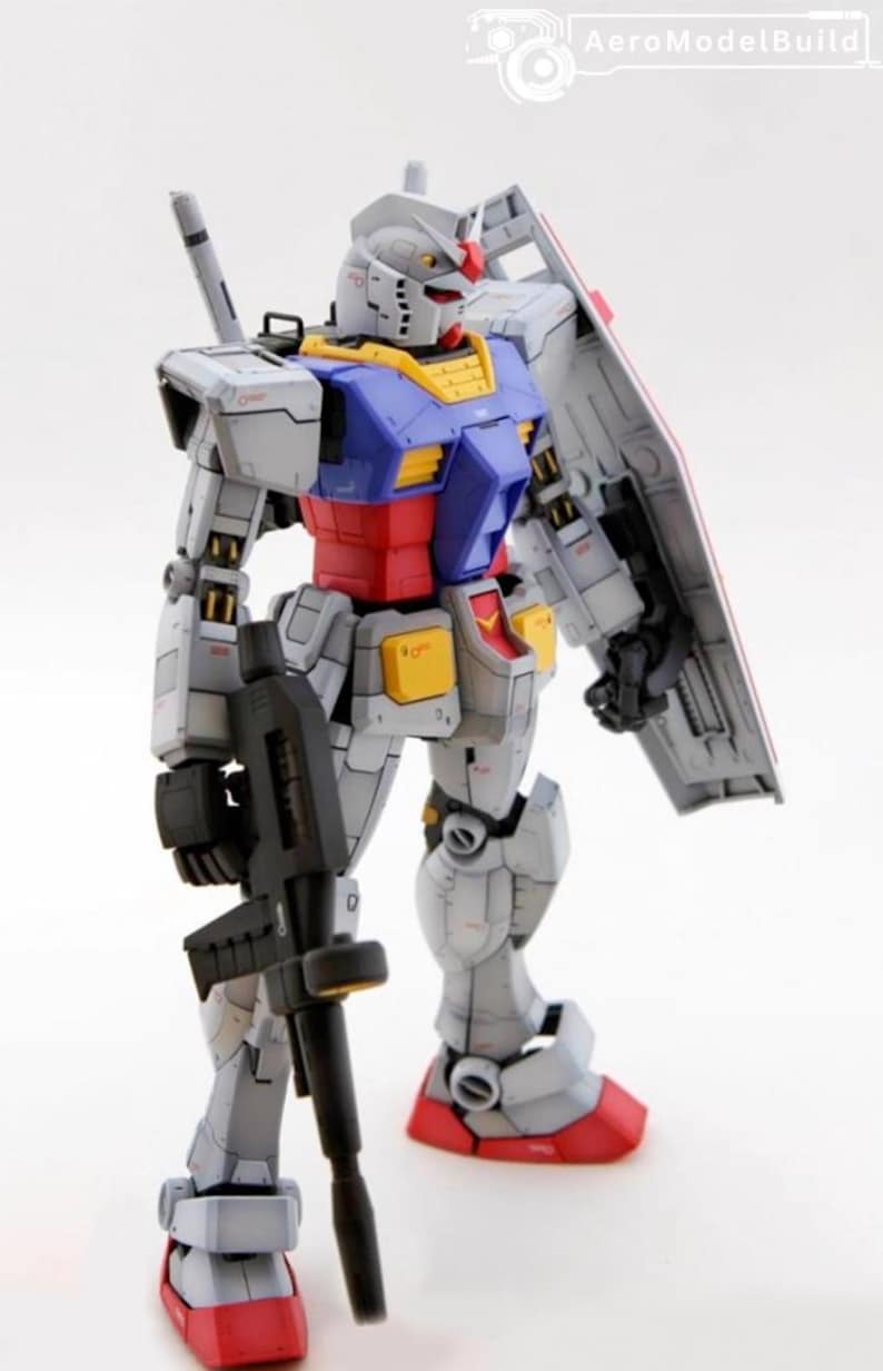 May include: A detailed model of a Gundam robot, primarily gray with blue, red, and yellow accents. It holds a black rifle and a shield. The model is standing upright, showcasing its intricate design and mechanical details.