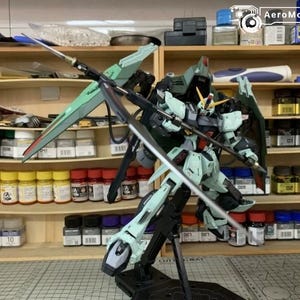 Puede incluir: Maqueta detallada de un robot Gundam, principalmente en tonos verdes, grises y negros, con grandes alas y una espada. El modelo est&aacute; posado sobre un soporte negro, con un taller de fondo.