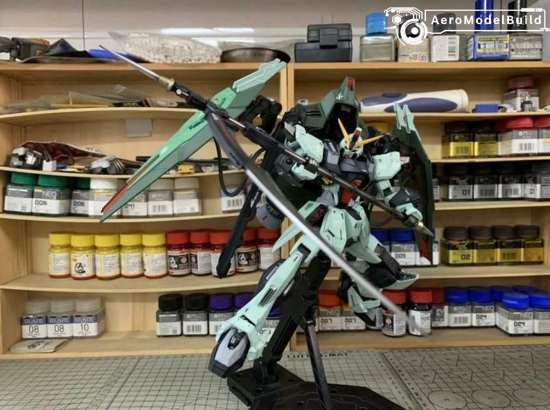 Puede incluir: Maqueta detallada de un robot Gundam, principalmente en tonos verdes, grises y negros, con grandes alas y una espada. El modelo est&aacute; posado sobre un soporte negro, con un taller de fondo.