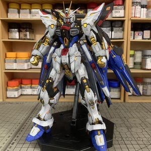 Puede incluir: Un detallado kit de modelo de un robot Gundam, con una combinaci&oacute;n de colores blanco, azul, negro y dorado. El modelo est&aacute; posado sobre un soporte negro, mostrando intrincados detalles mec&aacute;nicos y elementos de dise&ntilde;o. Incluye el texto "ArrowModelBuild".