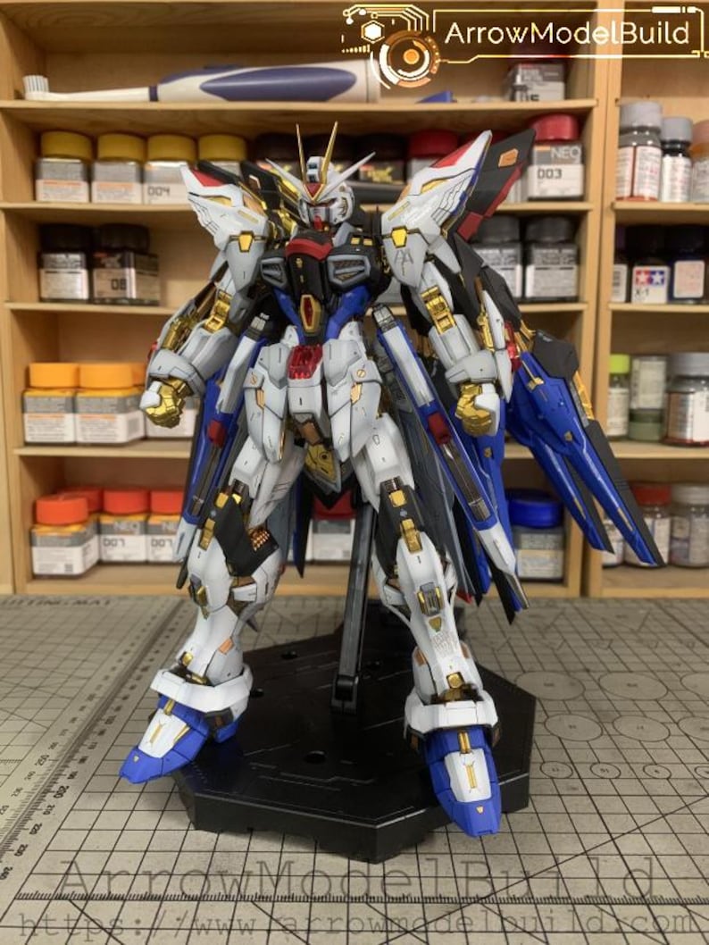 Puede incluir: Un detallado kit de modelo de un robot Gundam, con una combinaci&oacute;n de colores blanco, azul, negro y dorado. El modelo est&aacute; posado sobre un soporte negro, mostrando intrincados detalles mec&aacute;nicos y elementos de dise&ntilde;o. Incluye el texto "ArrowModelBuild".